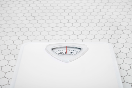 A white analog bathroom scale sitting on a bright floor made of white hexagonal tiles.