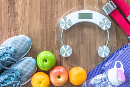 Running shoes, fruit, water bottle, weights, yoga mat, and scale sitting on a hardwood floor, depicting someone living an active and healthy lifestyle day-to-day. | Soza Weight Loss