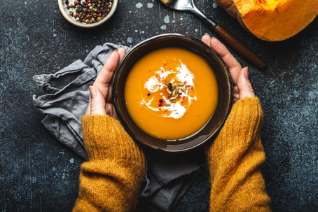 Woman sitting down to eat pumpkin soup for dinner on an autumn evening. She is wearing a yellow sweater and warming her hands on the bowl. | Soza Weight Loss