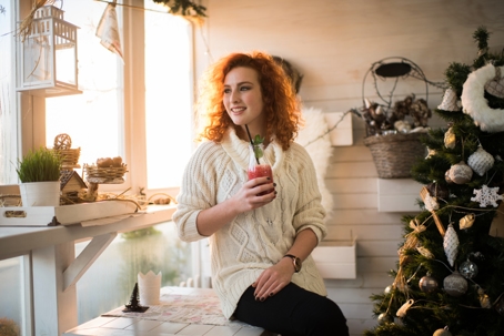 Woman smiling and looking out the window while drinking a healthy berry smoothie by a decorated tree over the holidays. | Soza Weight Loss