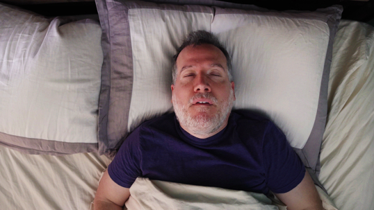 An overhead view of a middle-aged man sleeping on his back in bed with his mouth slightly open, a common sign of snoring or sleep apnea.