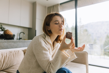 Woman sitting on a couch enjoying a piece of chocolate, showing how comfort foods can influence mood and cravings.