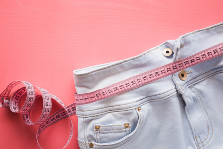 Tape measure wrapped around jeans on a pink background, representing weight changes and daily fluctuations on the scale.