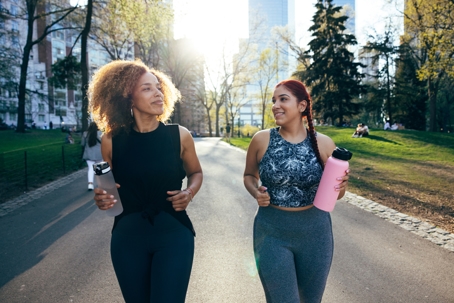 Two women happily jogging through a city park on a nice day. The sun is shining and they both have water bottles in hand. | Soza Weight Loss